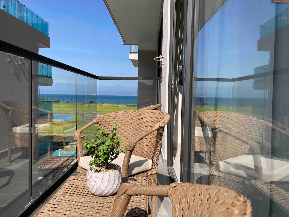A balcony area is shown, featuring two woven chairs positioned by a small potted plant. The glass rail affords views of the ocean and surrounding landscape, while large windows reflect the bright sky and sunlight.