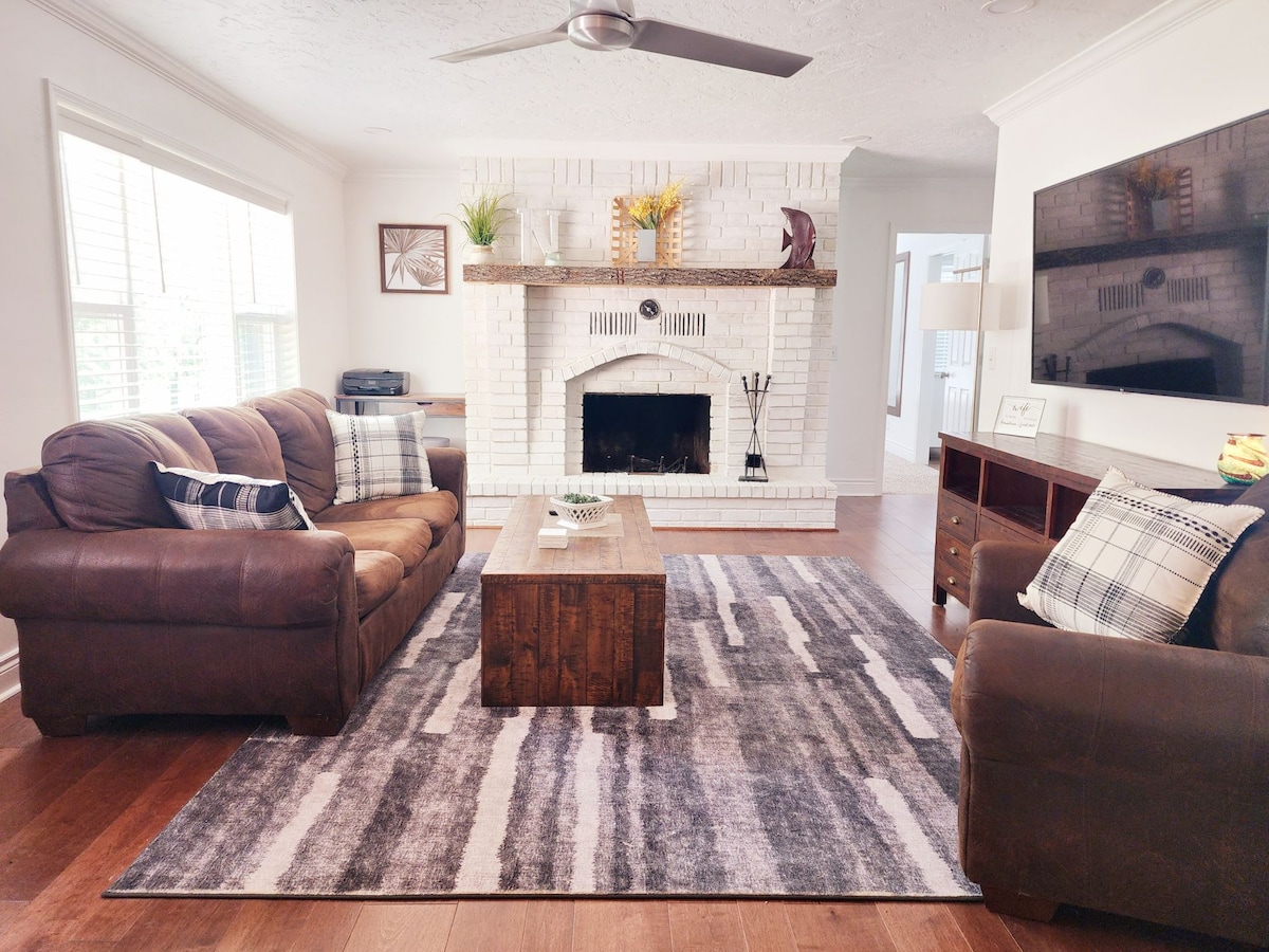 A cozy living room is highlighted by a brown leather couch and matching chair, arranged around a wooden coffee table. A textured area rug covers the floor, complementing the white-brick fireplace. Natural light enters through the windows, enhancing the space's inviting feel.