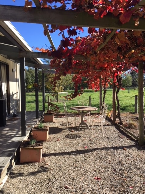 An outdoor seating area is shaded by vibrant autumn leaves, featuring a round table accompanied by decorative chairs. The gravel path leads to a lawn framed by rustic fencing, creating a serene rural ambiance.