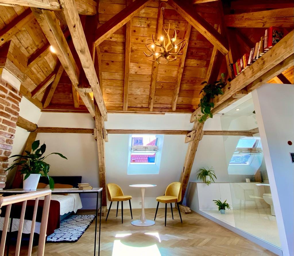 The studio features a high wooden ceiling with exposed beams, enhancing its cozy character. Two yellow chairs are paired with a small round table, while a sleek glass partition allows natural light to fill the space. A bed is visible in the background, creating a welcoming atmosphere.