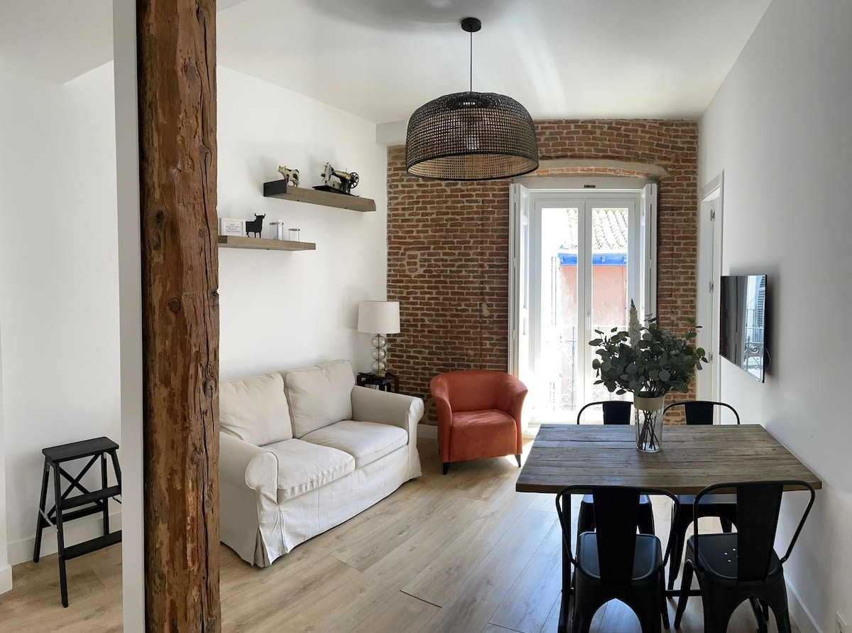 A comfortable living area features a white sofa paired with an orange armchair. A wooden dining table is surrounded by black chairs. Exposed brick walls add character, while large doors lead to a balcony, allowing natural light to brighten the space.