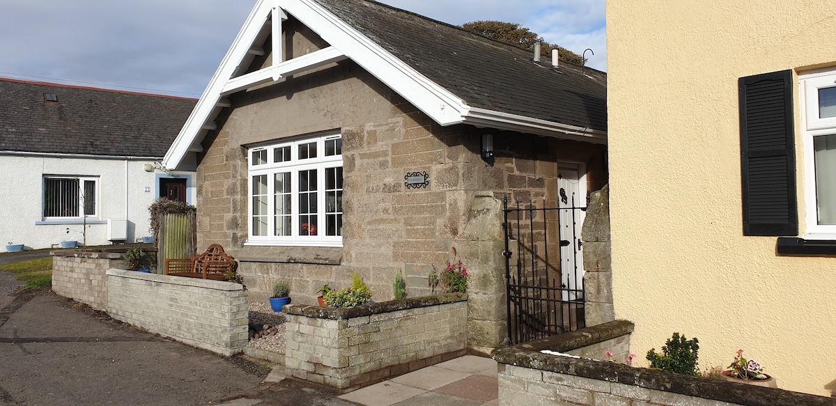 The charming seafront cottage displays a stone facade with decorative windows framed by dark shutters. A low stone wall encircles a neatly landscaped garden, featuring small plants and a seating area. The entrance is marked by a black gate, leading to a welcoming front door.