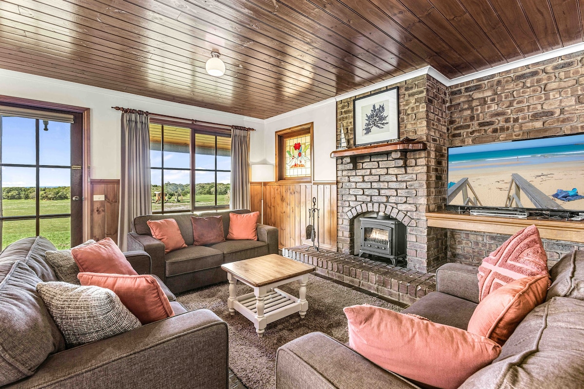 A comfortable living area features two sofas with loose cushions, arranged around a central wooden coffee table. Natural light enters through large windows, illuminating the room's brick fireplace and rustic decor, which includes a framed artwork and a flat-screen television mounted on the wall.