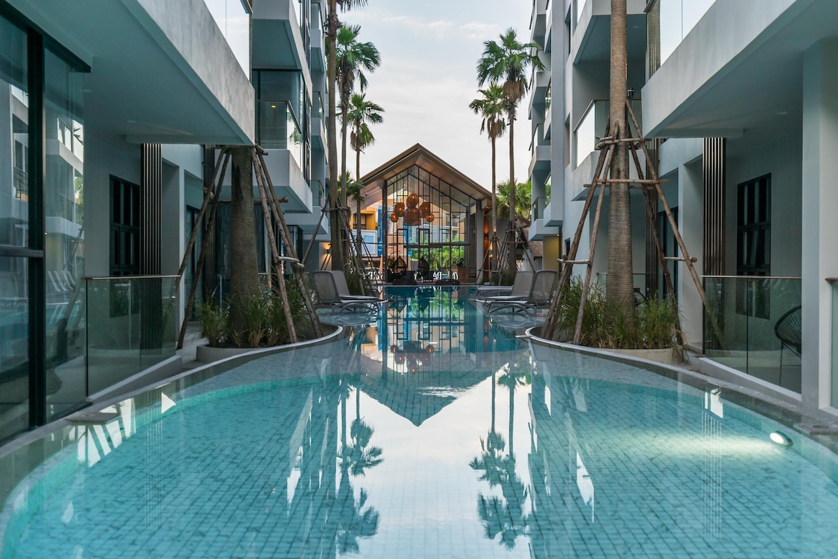 A serene view of the ground floor long swimming pool is showcased, flanked by palm trees and modern architectural elements. Lounge chairs are positioned alongside the water, reflecting the glass-walled communal space in the distance.