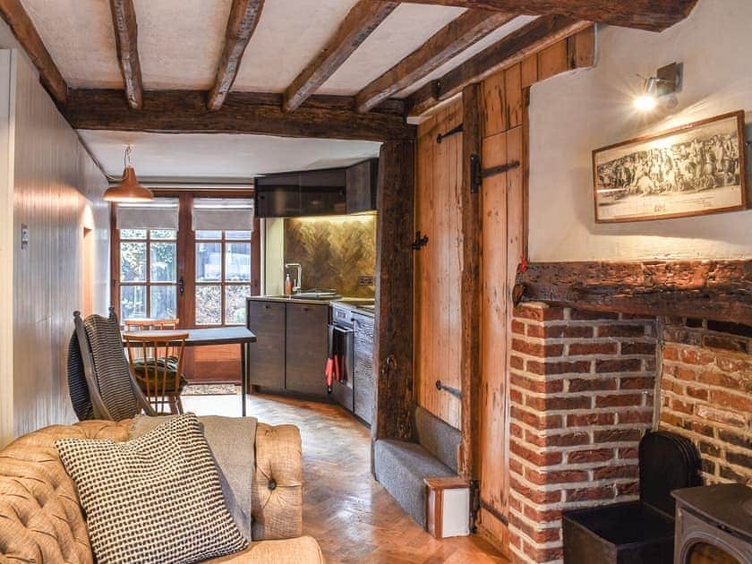An open-plan living area is illuminated by natural light from large windows. Exposed wooden beams add warmth to the space, featuring a comfortable chesterfield sofa and a traditional inglenook fireplace. The modern kitchen, equipped with integrated appliances, is seamlessly integrated into the cozy design.