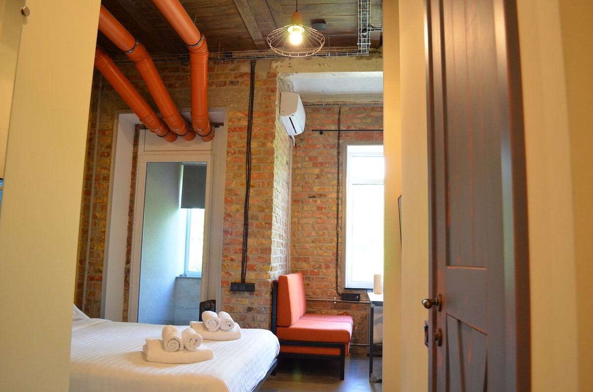 A studio space features exposed brick walls and warm wooden accents. A spacious bed is complemented by neatly arranged towels. A corner seating area with vibrant orange upholstery adds a pop of color, while a window allows natural light to fill the room.
