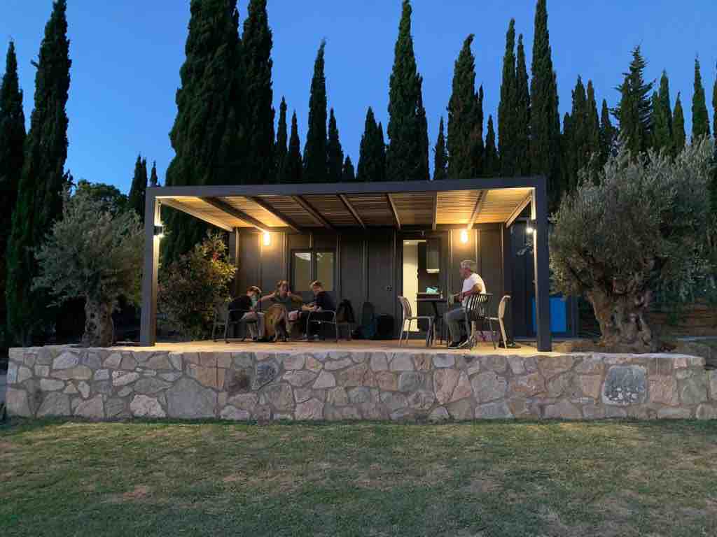 An outdoor terrace is presented with a stone base and a covered seating area. Four individuals are gathered, with a table and chairs arranged under the shelter, surrounded by tall, green trees. Soft lighting enhances the evening ambiance.