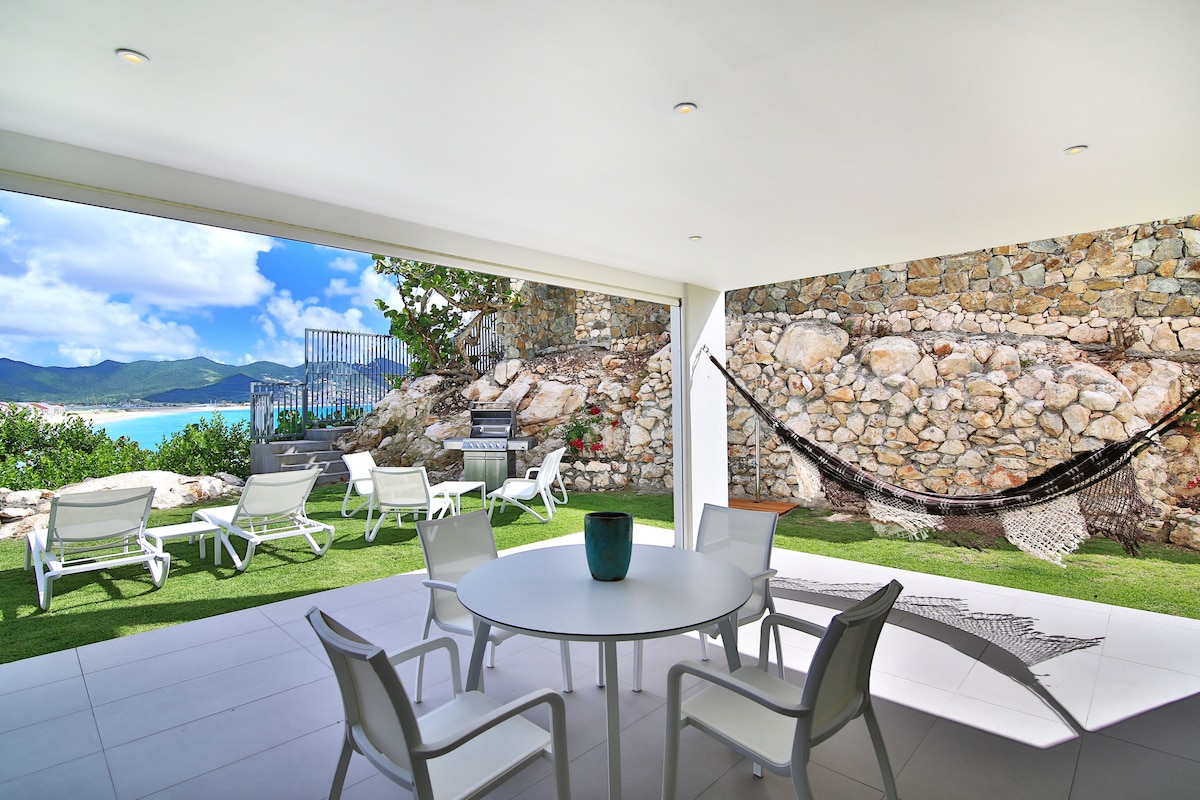 An outdoor patio area features a round dining table surrounded by white chairs. A hammock is positioned nearby, with a view of lush greenery and the ocean beyond. The patio is accented by stone walls and provides access to a peaceful, natural setting.