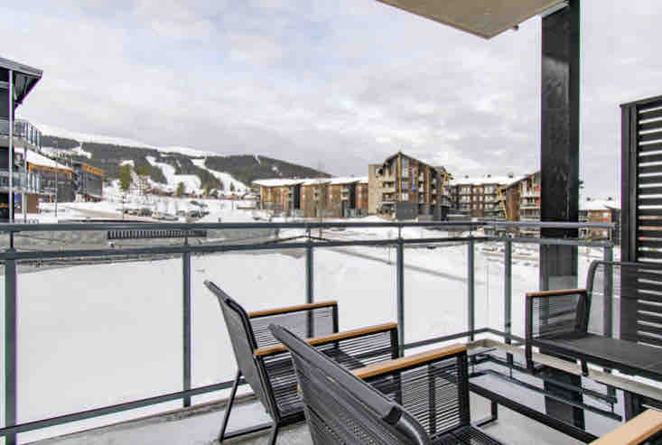 A balcony offers a view of the surrounding snow-covered landscape and buildings in the distance. Two black lounge chairs with wooden accents are positioned on the balcony, providing a place to relax and enjoy the winter scenery.