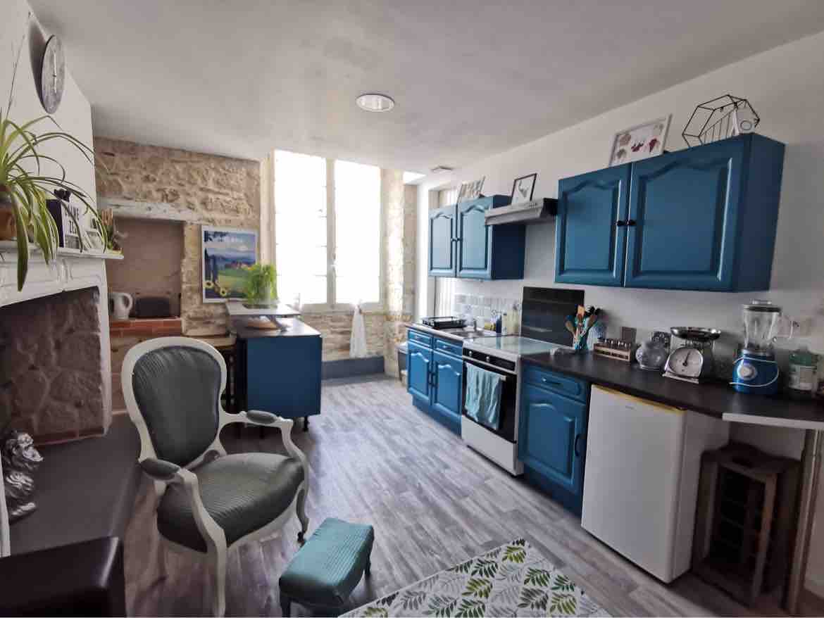 A kitchen area is highlighted by blue cabinetry and stone walls. Natural light enters through large windows, illuminating modern appliances, including a stove and refrigerator. A small round table is paired with a white chair, while decorative items and plants add character to the space.