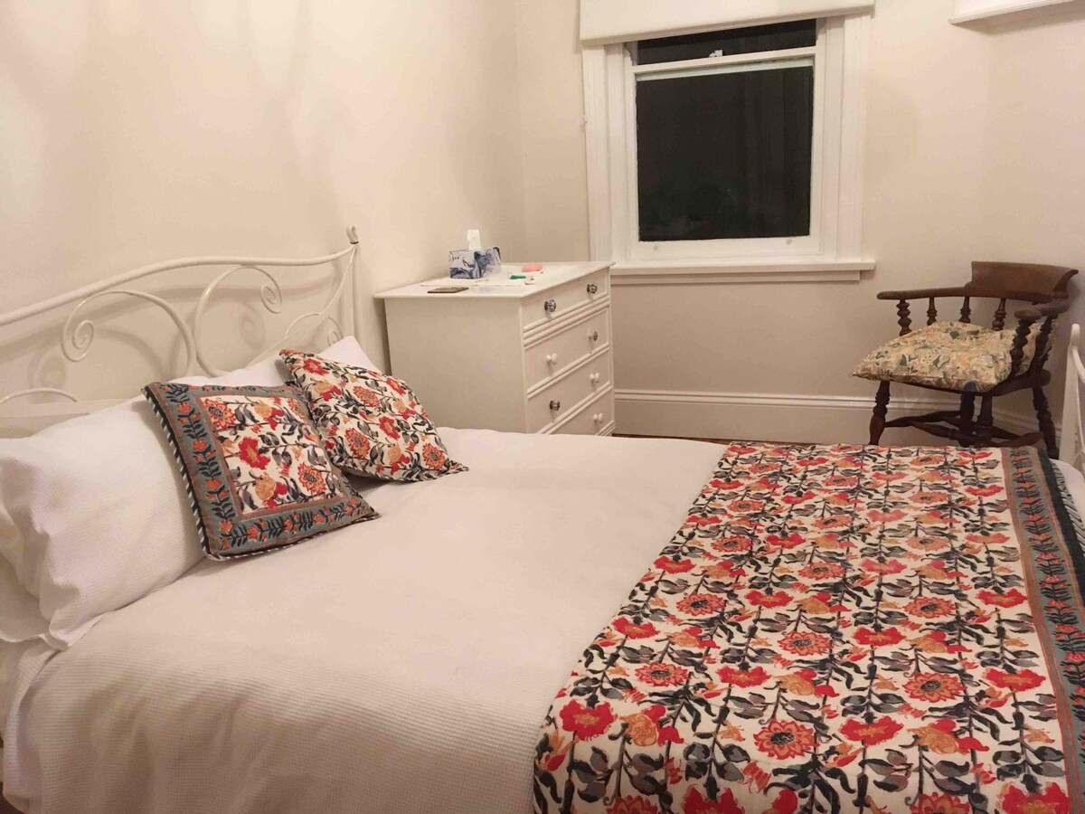 A cozy bedroom is presented with a spacious bed adorned with a floral patterned blanket and matching decorative pillows. A white dresser is positioned nearby, complementing the light walls. Natural light filters through the window, illuminating a wooden chair with a patterned cushion.