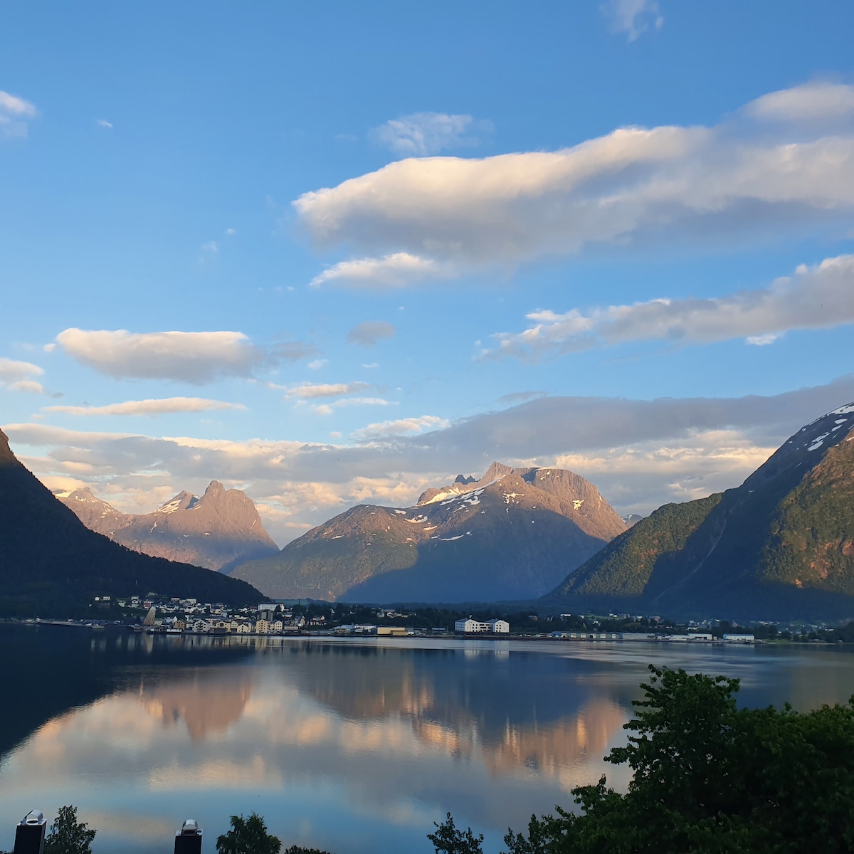 A panoramic view of calm waters reflects surrounding mountains under a partly cloudy sky. The landscape features lush greenery in the foreground, with distant peaks rising majestically. The tranquil scene emphasizes the serene beauty of the area.