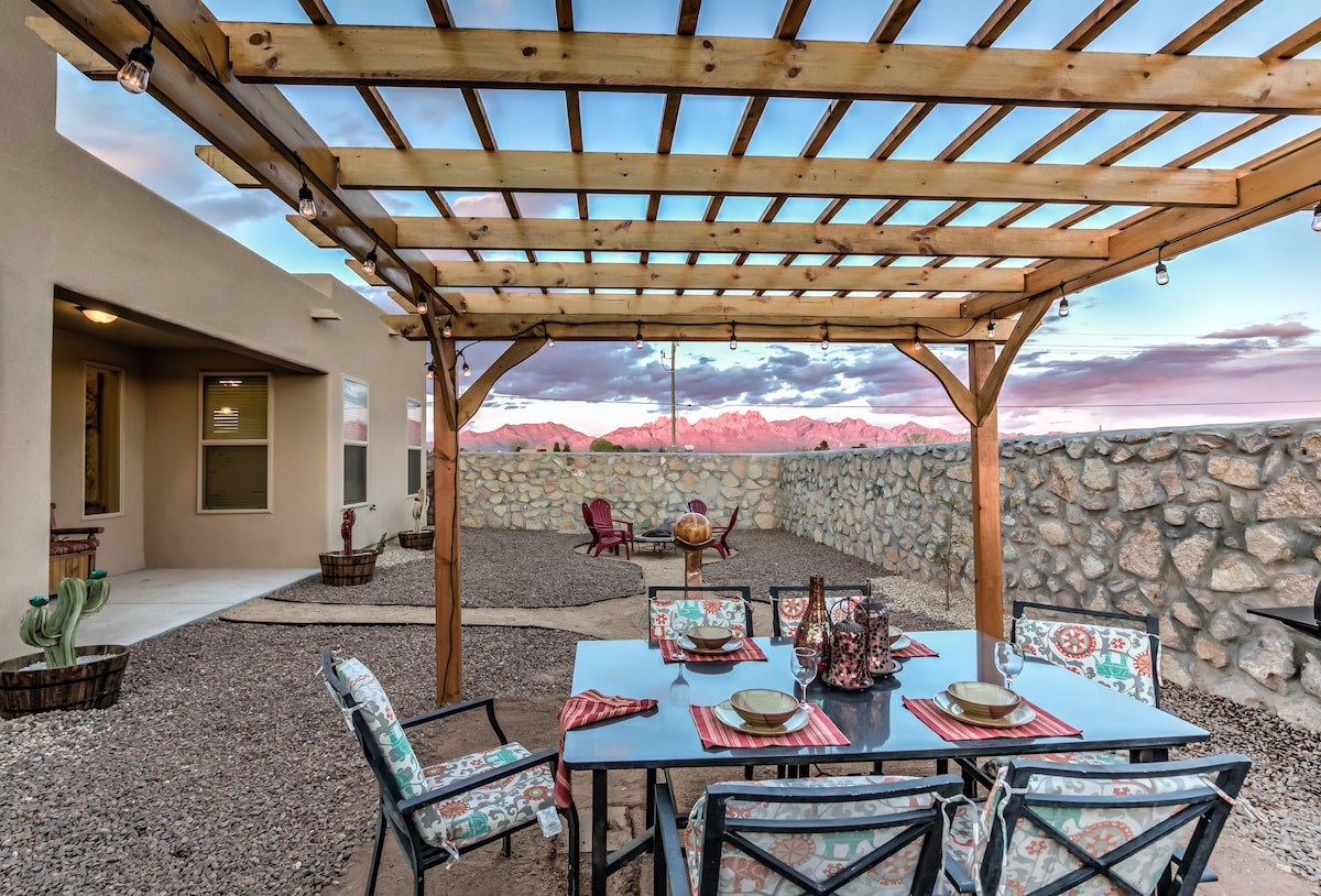 An outdoor dining area is framed by a wood pergola, featuring a black table set with plates and utensils. Two decorative chairs and a fire pit are visible, with gravel grounding the space. The Organ Mountains are seen in the background beneath a colorful sky.