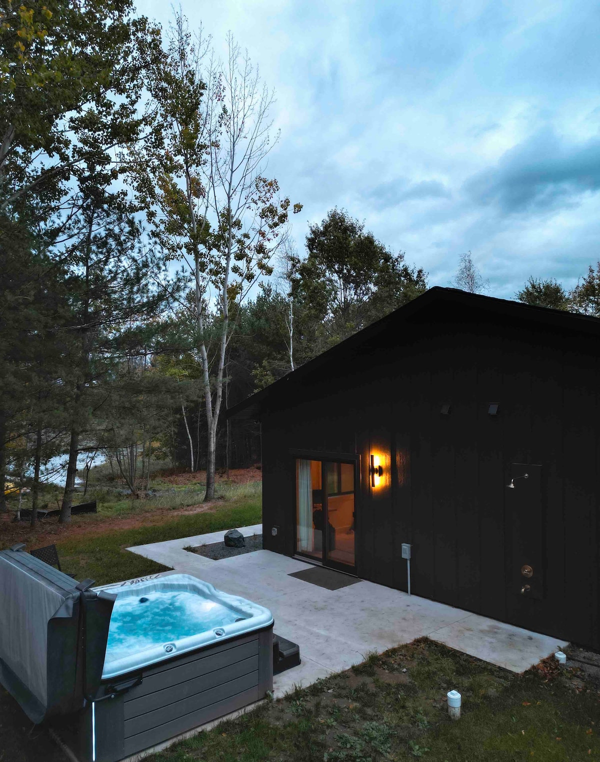 An outdoor area showcases a hot tub set on a concrete patio, nestled among trees. The dark exterior of the cabin provides a modern contrast to the natural surroundings. Soft evening light emanates from the cabin, enhancing the tranquil atmosphere.