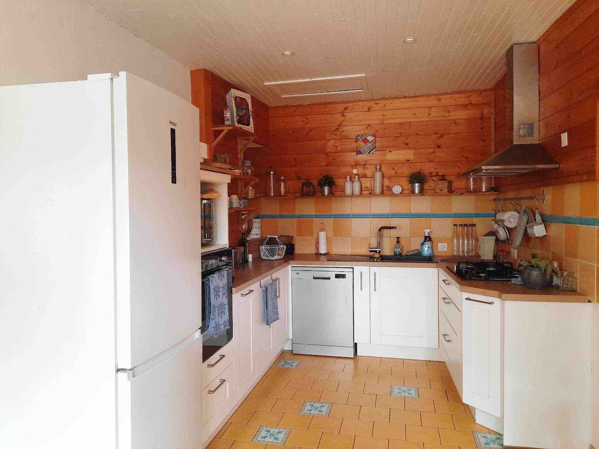 A well-equipped kitchen is showcased, featuring a large refrigerator, a dishwasher, and a gas stove. Wooden cabinetry provides a warm feel, complemented by tiled countertops. Shelving displays kitchenware, while abundant natural light illuminates the space.