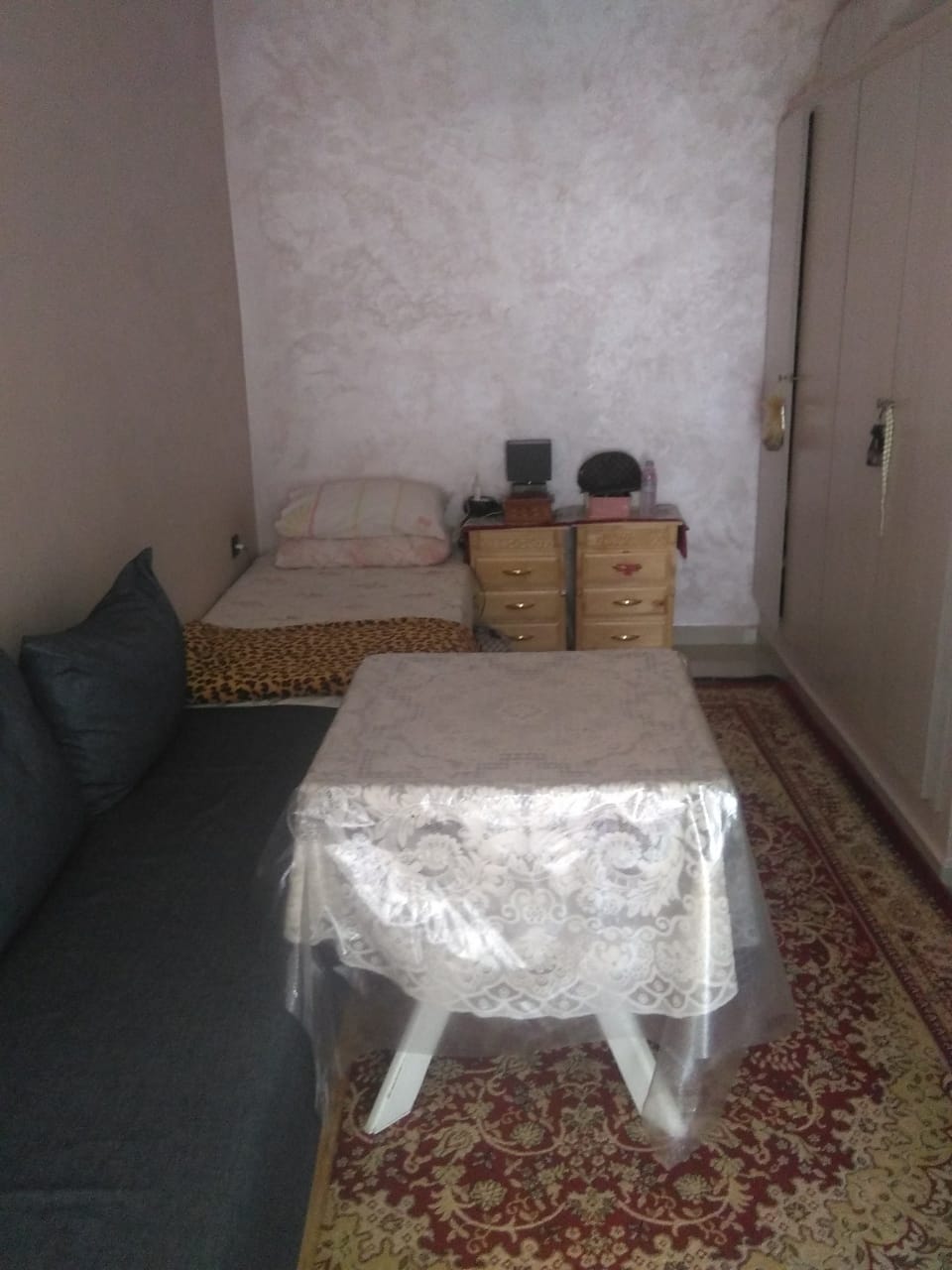 The room contains a single bed and a sofa bed, with a small table in front. A dresser with multiple drawers is visible beside the bed. The floor features a decorative rug, and the walls are finished in a light tone.