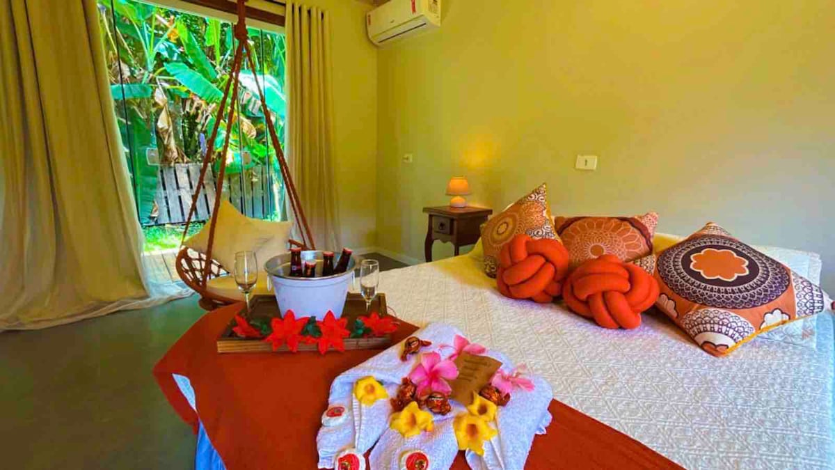 A spacious room is shown with a cozy bed adorned with decorative pillows. A small table with glasses and a bowl is placed on a blanket at the foot of the bed. Large sliding doors open to a lush outdoor area, adding natural light.