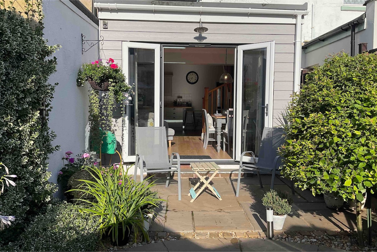 The garden area features a paved patio with two folding chairs and a small table. Lush greenery surrounds the space, including potted plants and hanging flower baskets, creating a serene outdoor setting suitable for relaxation.