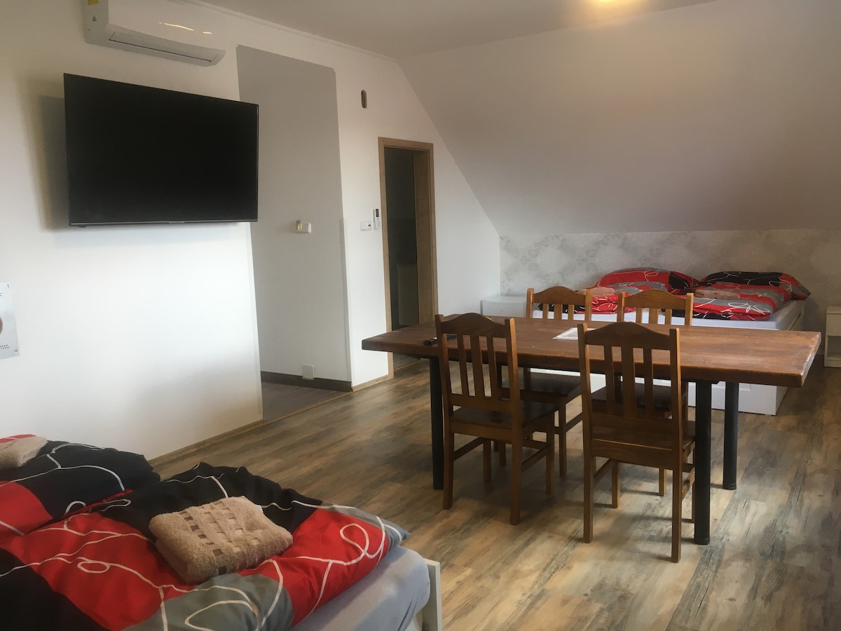 A bright room is furnished with two double beds and a wooden dining table surrounded by four chairs. A television is mounted on the wall opposite the seating area. Natural light streams in, highlighting the warm tones of the wooden flooring.