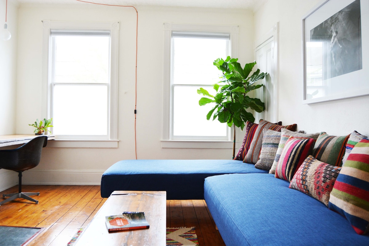 A bright living area features a blue sectional sofa adorned with colorful patterned cushions. Natural light enters through two windows, creating an open atmosphere. A wooden coffee table rests in front of the sofa, and a potted plant adds greenery to the space.