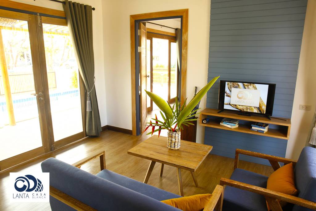 A seating area is displayed with comfortable blue armchairs positioned around a wooden coffee table. A flat-screen TV is mounted on a wooden shelf against a wall, and large glass doors provide access to the outdoor space, allowing natural light to fill the room.