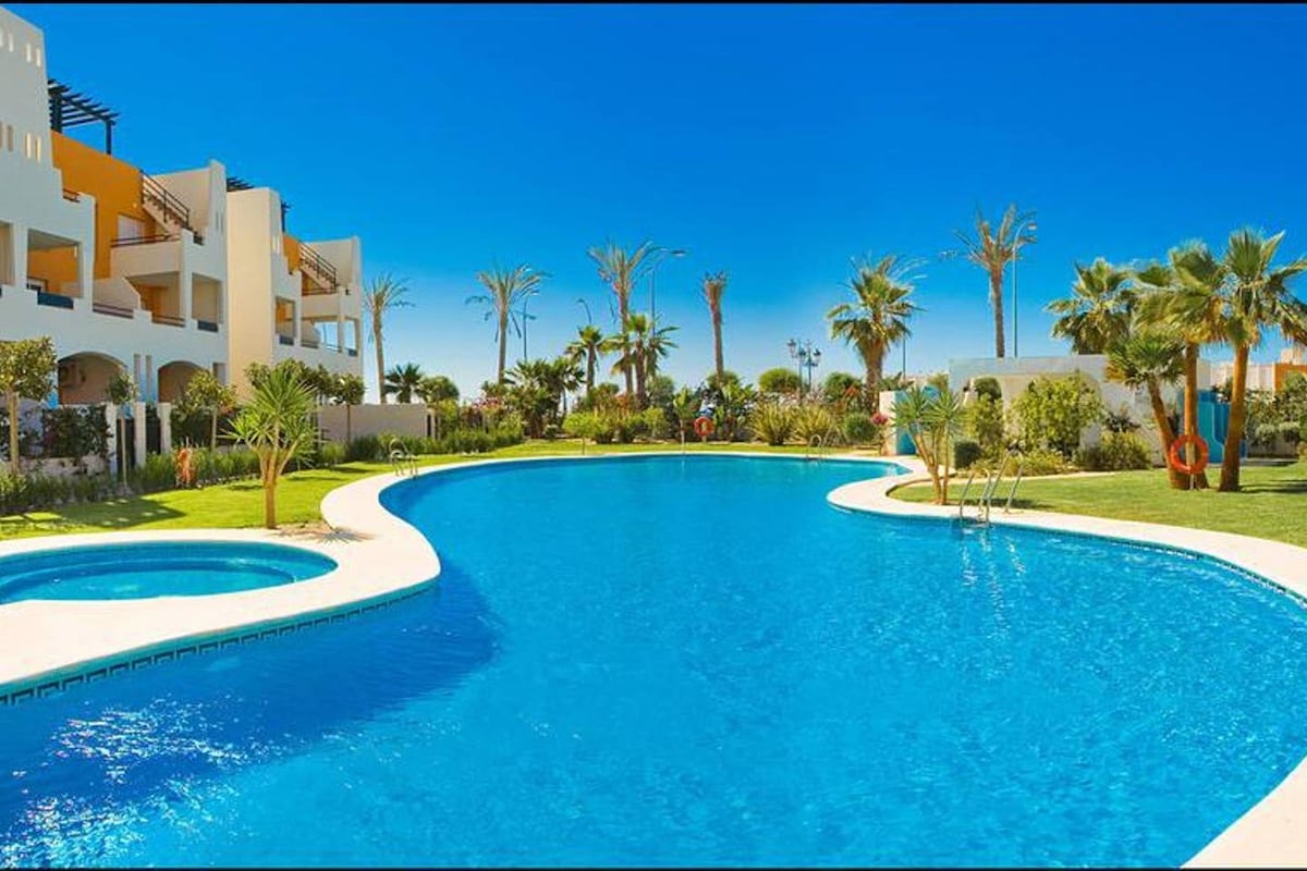 A large, clear swimming pool is surrounded by well-maintained gardens featuring palm trees and various green plants. The pool's reflective surface mirrors the bright blue sky, while lounge areas are visible nearby, providing ample space for relaxation.