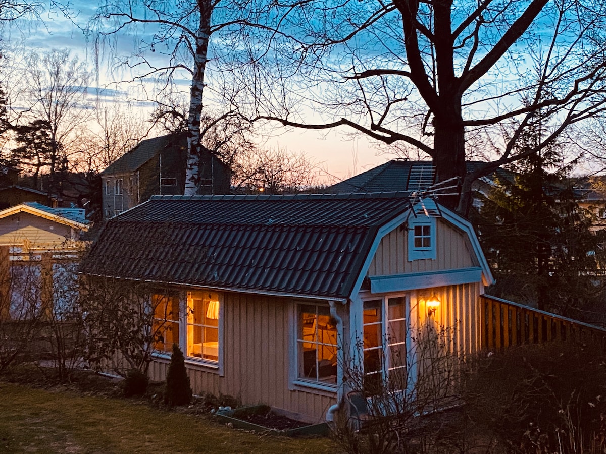 A small cottage is illuminated by warm lights, standing amidst a serene outdoor setting. Large windows reflect the soft hues of the sunset sky. Nearby trees provide a natural backdrop, enhancing the tranquil atmosphere of the garden area.