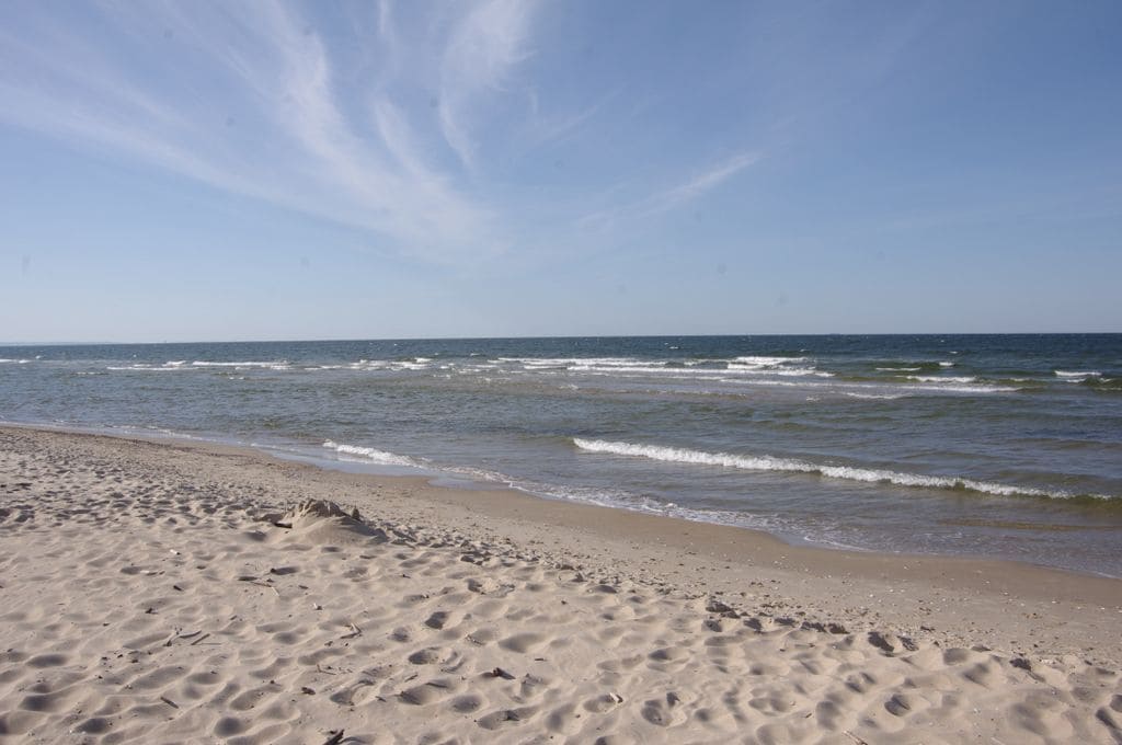 A wide and tranquil beach is seen, with soft sand extending toward gentle waves lapping at the shore. The horizon features a clear blue sky with light clouds, creating a serene atmosphere. The absence of crowds enhances the peaceful surroundings.