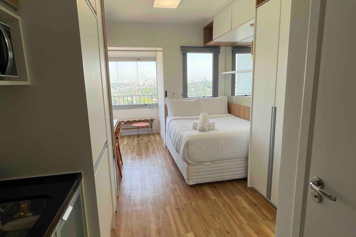 A compact studio apartment is displayed, featuring a spacious bed with white linens and a towel folded into a decorative shape. A large window offers city views, while built-in cabinets provide storage. Natural light enhances the warm wood flooring throughout the space.