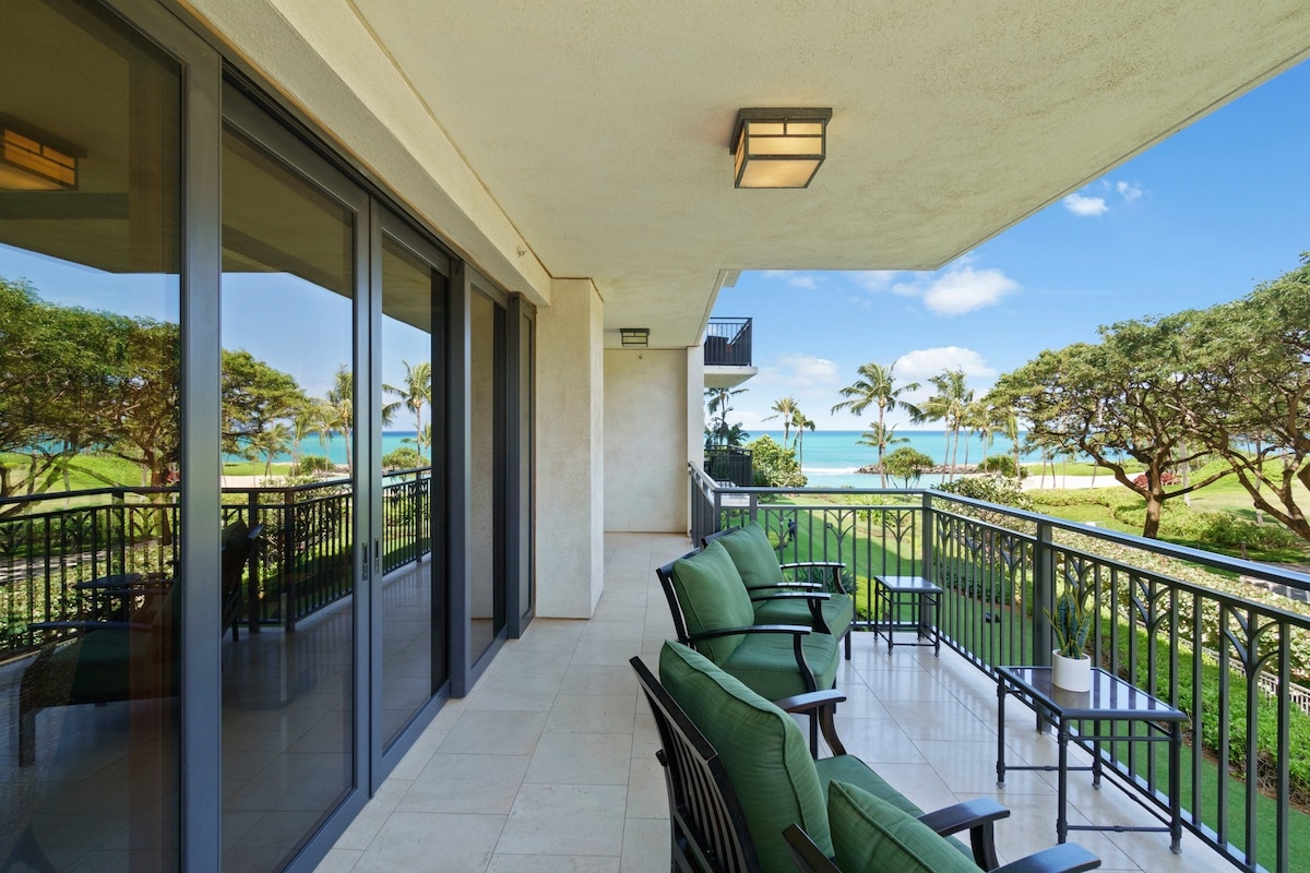 A spacious balcony features six comfortable chairs arranged around a small table, overlooking lush greenery and a glimpse of the ocean. Large glass doors reflect natural light, enhancing the open feel of the outdoor space. Palm trees sway gently in the breeze.