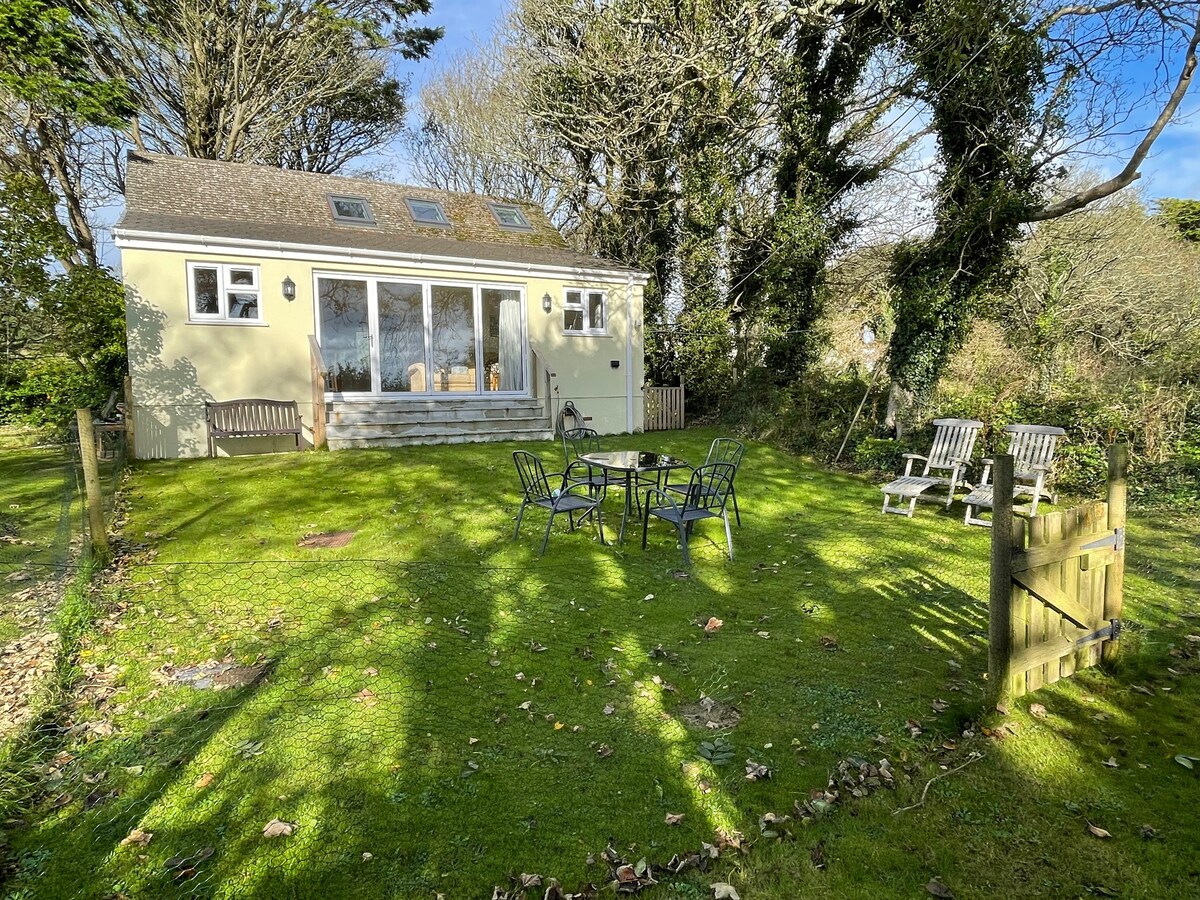A tranquil exterior view of the Lodge is presented, featuring a grassy yard surrounded by trees. The structure showcases large bi-fold doors, allowing natural light to fill the interior. Outdoor furniture, including a table with chairs and two loungers, is arranged on the lawn.