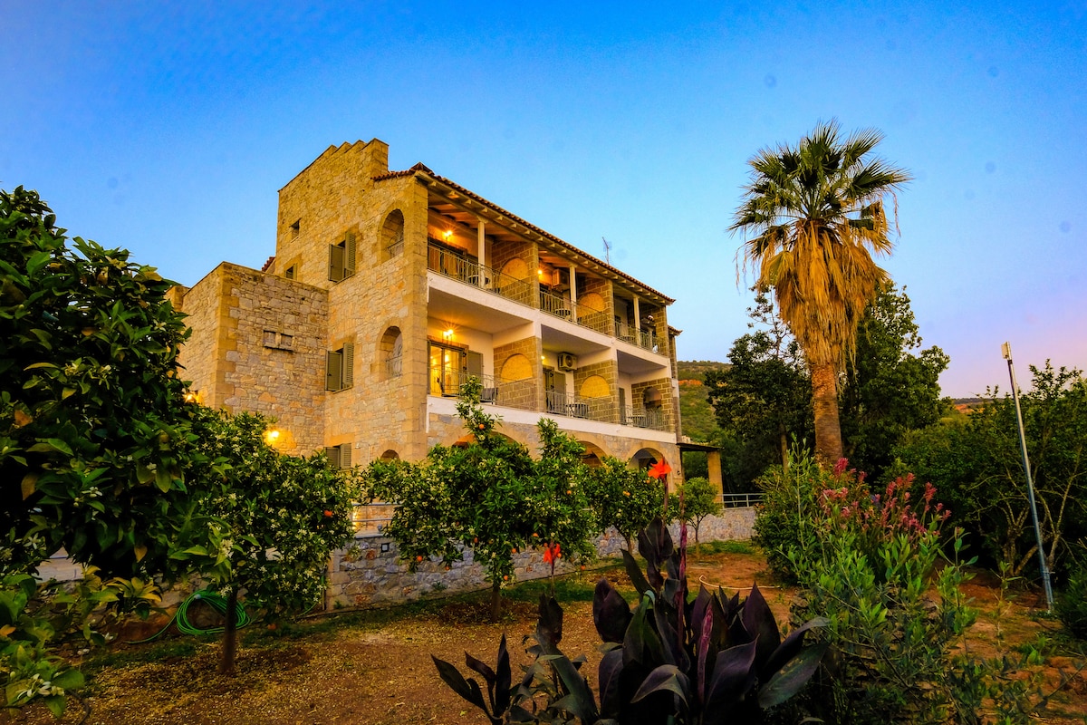 The traditional two-story building is surrounded by a lush garden featuring a variety of plants and trees. Large covered verandas provide outdoor seating options, while warm evening light illuminates the stone facade. Palm and flowering plants can be seen in the garden area.