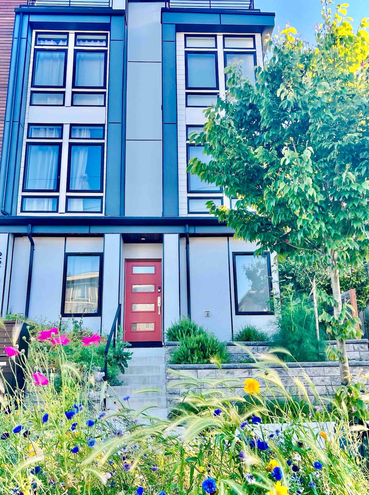 The exterior of the townhome displays a modern façade with large windows. A vibrant red front door stands out against the neutral colors of the walls. An array of colorful flowers and greenery adorns the entrance, complemented by clean stone steps leading to the door.