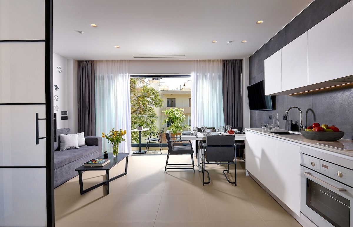 A spacious living area features a comfortable sofa, a dining table set for four, and a fully equipped kitchen. Large windows allow natural light to fill the room, revealing a view of the small balcony and outdoor seating. Neutral tones create a calm environment.