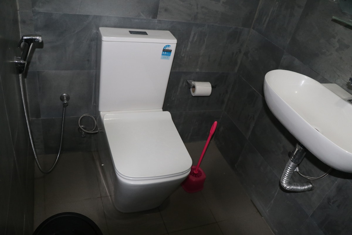 A modern bathroom is displayed, featuring a sleek white toilet and a small pink toilet brush nearby. A compact washbasin is mounted on the wall, with plumbing visible underneath. Grey tiles cover the walls and floor, enhancing the minimalist design.