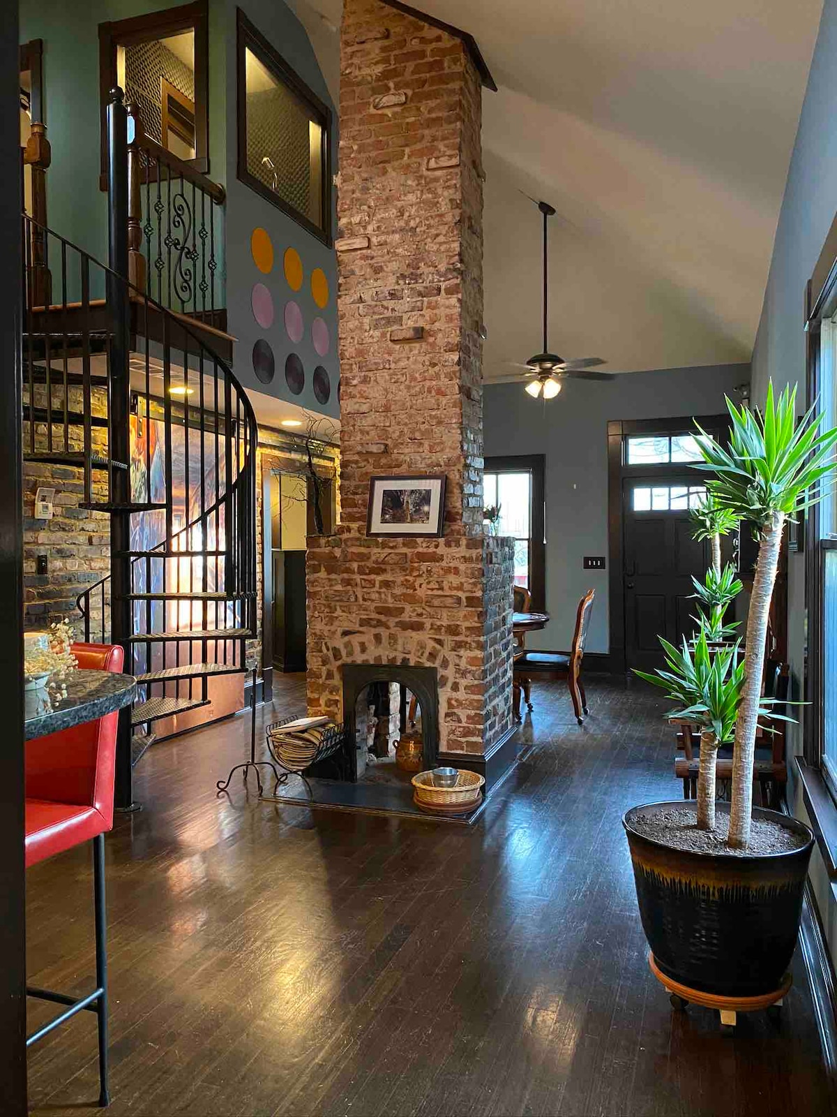 A warm entryway is complemented by a striking brick chimney, serving as a focal point. A spiral staircase leads to the upper level, while dining chairs are arranged near a window. Decorative plants and wall art add character to the space.
