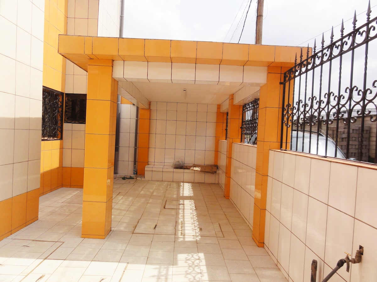 An outdoor parking area is characterized by tiled flooring and orange and white wall panels. The space features support columns and is surrounded by a decorative black iron fence. Natural light enters from above, casting patterns on the surface.