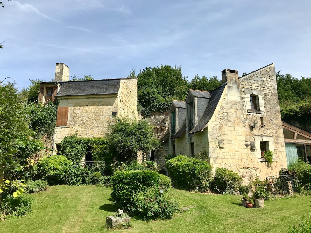 A charming stone property is set within a well-maintained garden, surrounded by lush greenery. The building features a variety of architectural elements, such as sloping roofs and windows, with the rustic stone exterior harmonizing with the natural landscape.