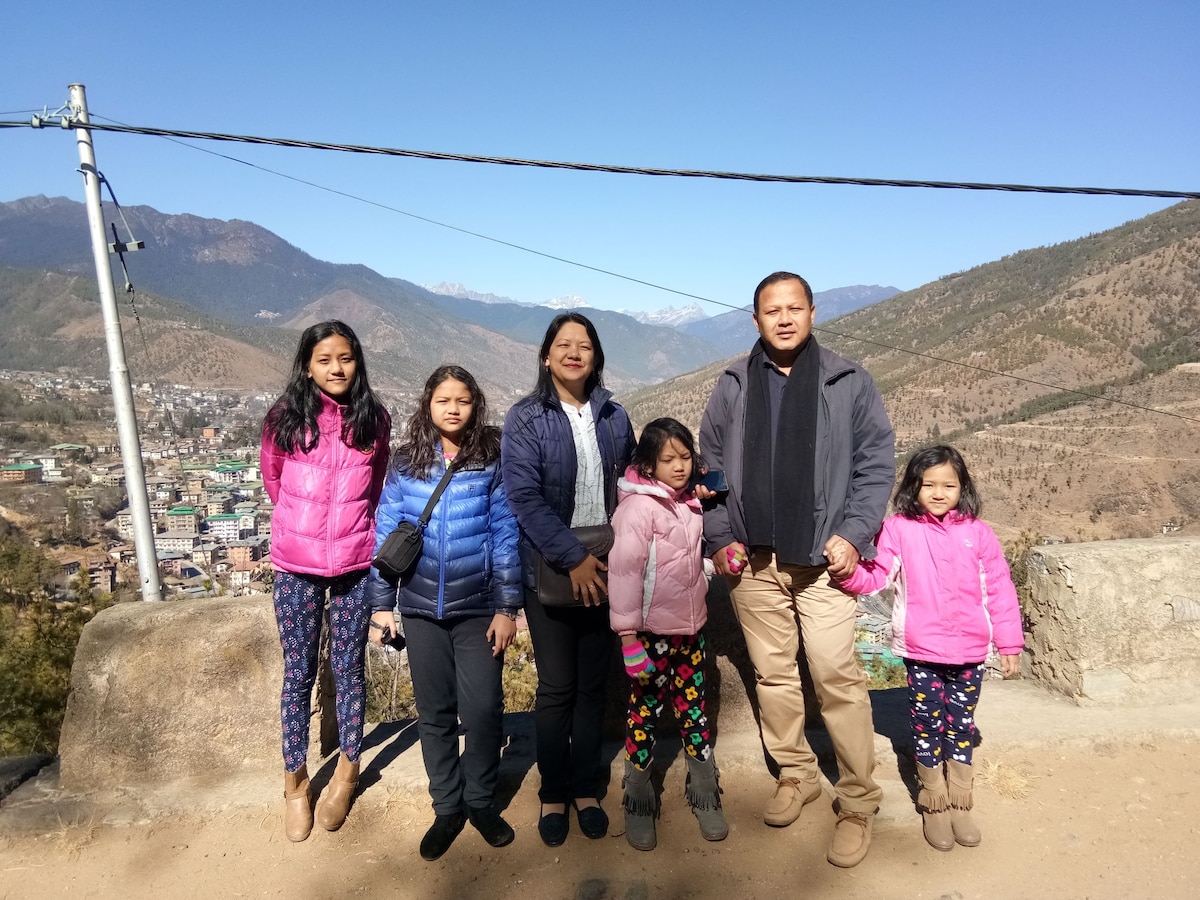 A group of five individuals stands on a rocky ledge, with a mountainous landscape in the background. The scene showcases a clear sky and expansive views of the hills and valleys. Each person is dressed in colorful winter jackets, adding vibrancy to the outdoor setting.