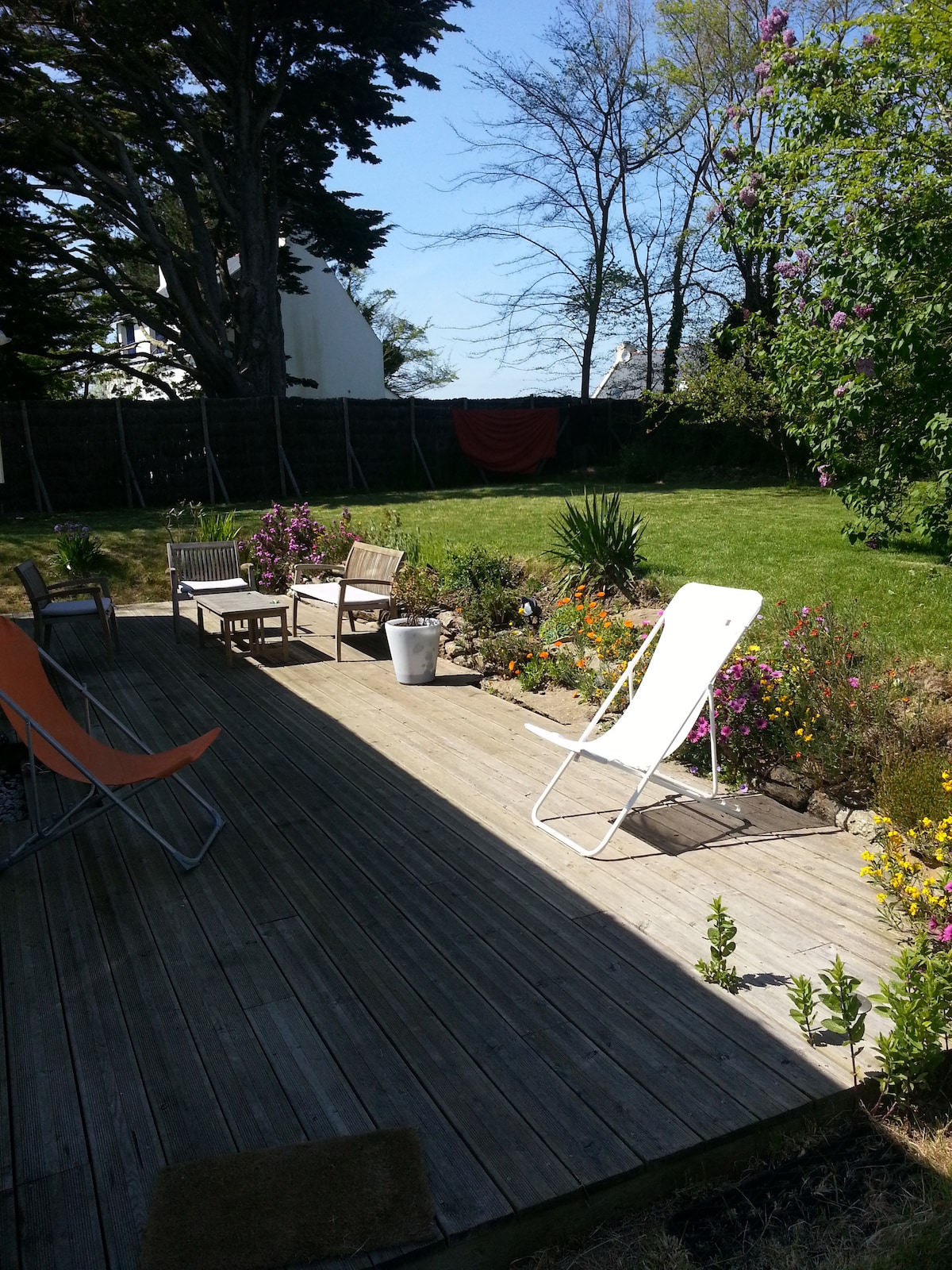 A wooden deck area features a mix of seating options, including a chaise lounge and two chairs. Colorful flowers and greenery are present along the edges, with a well-maintained lawn beyond. Bright sunlight creates contrasting areas of shade and light across the space.