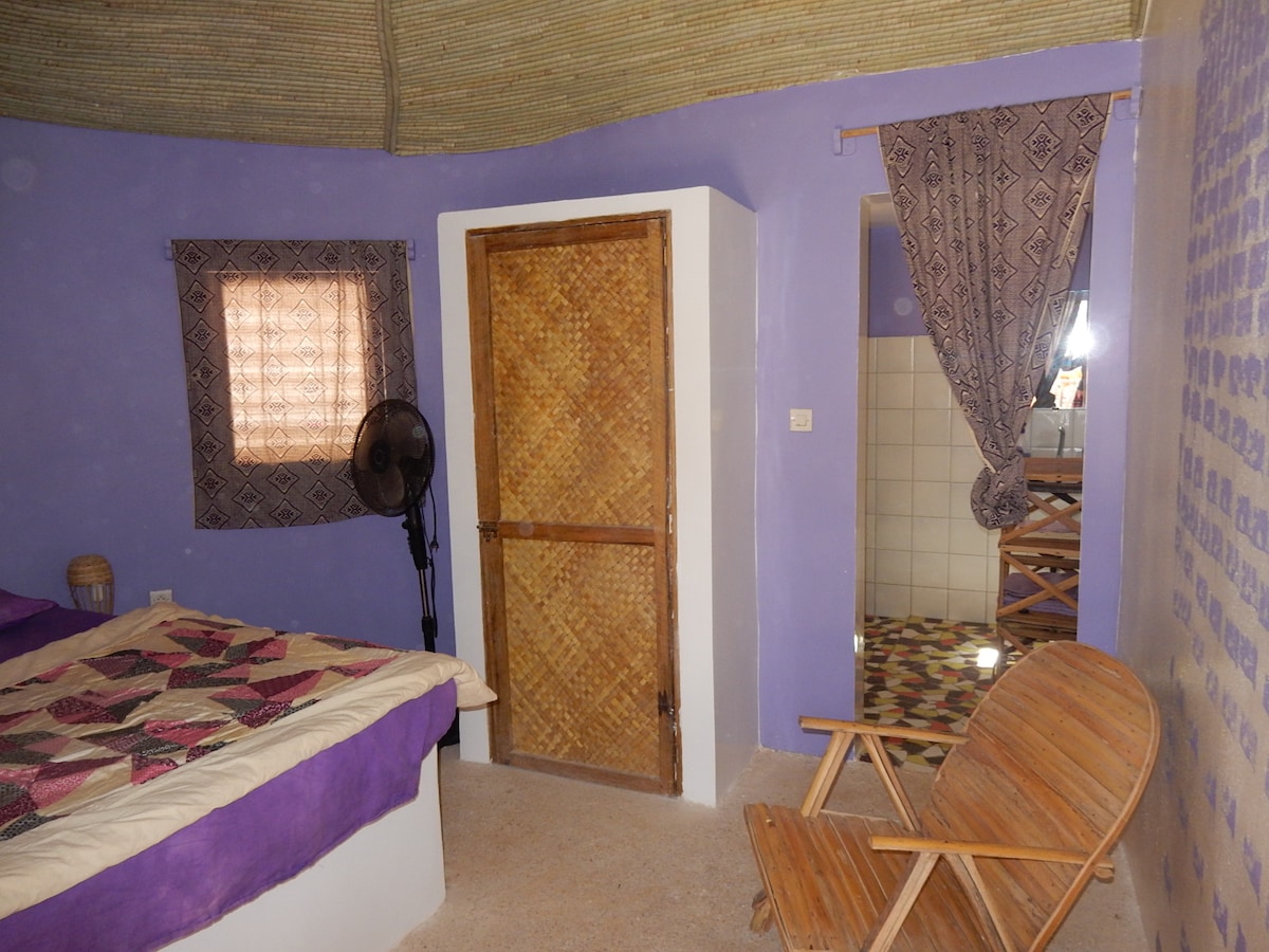 A serene sleeping area features a patchwork quilt covering a spacious bed. A wooden door leads to a bathroom area, visible through a doorway. Light enters from a nearby window, while a fan stands beside the bed. A wooden chair provides additional seating.