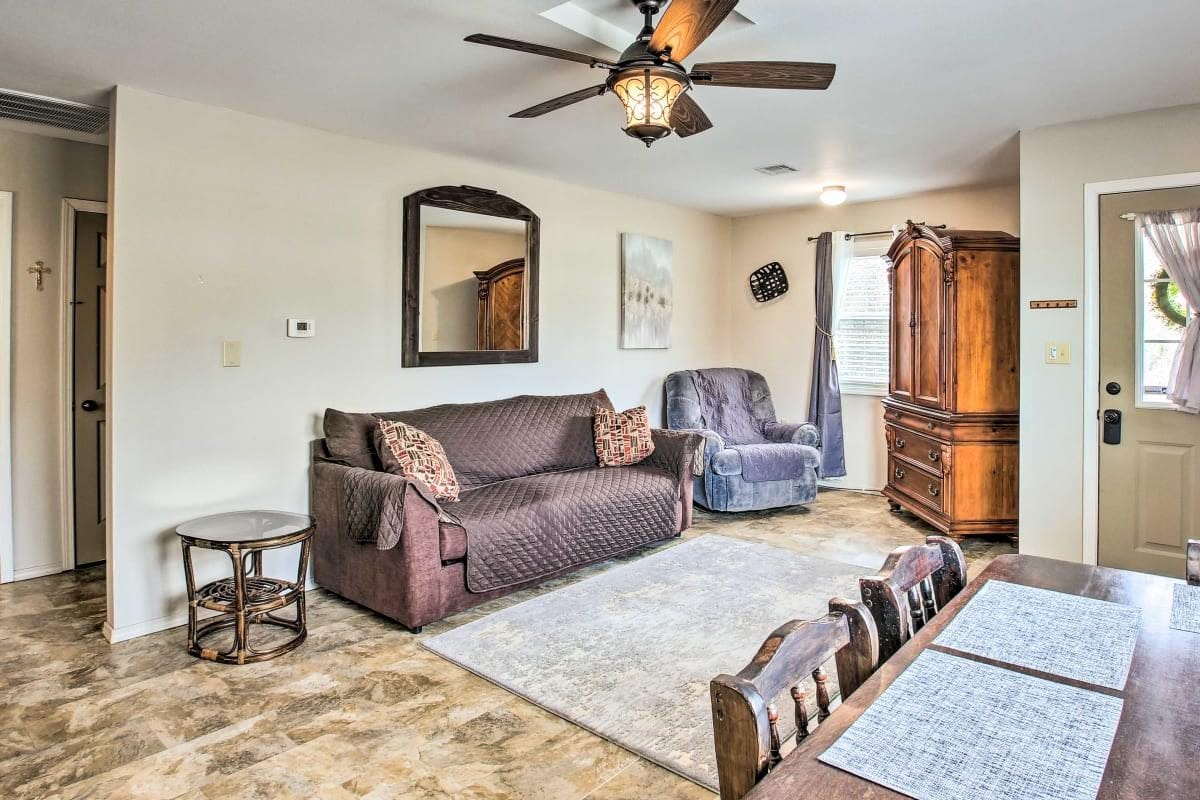 The living room is furnished with a plush sleeper sofa and a comfortable armchair. A large mirror is mounted on the wall, reflecting natural light. A wooden dining table is visible, accompanied by several rustic-style chairs. A ceiling fan gently circulates air in the space.