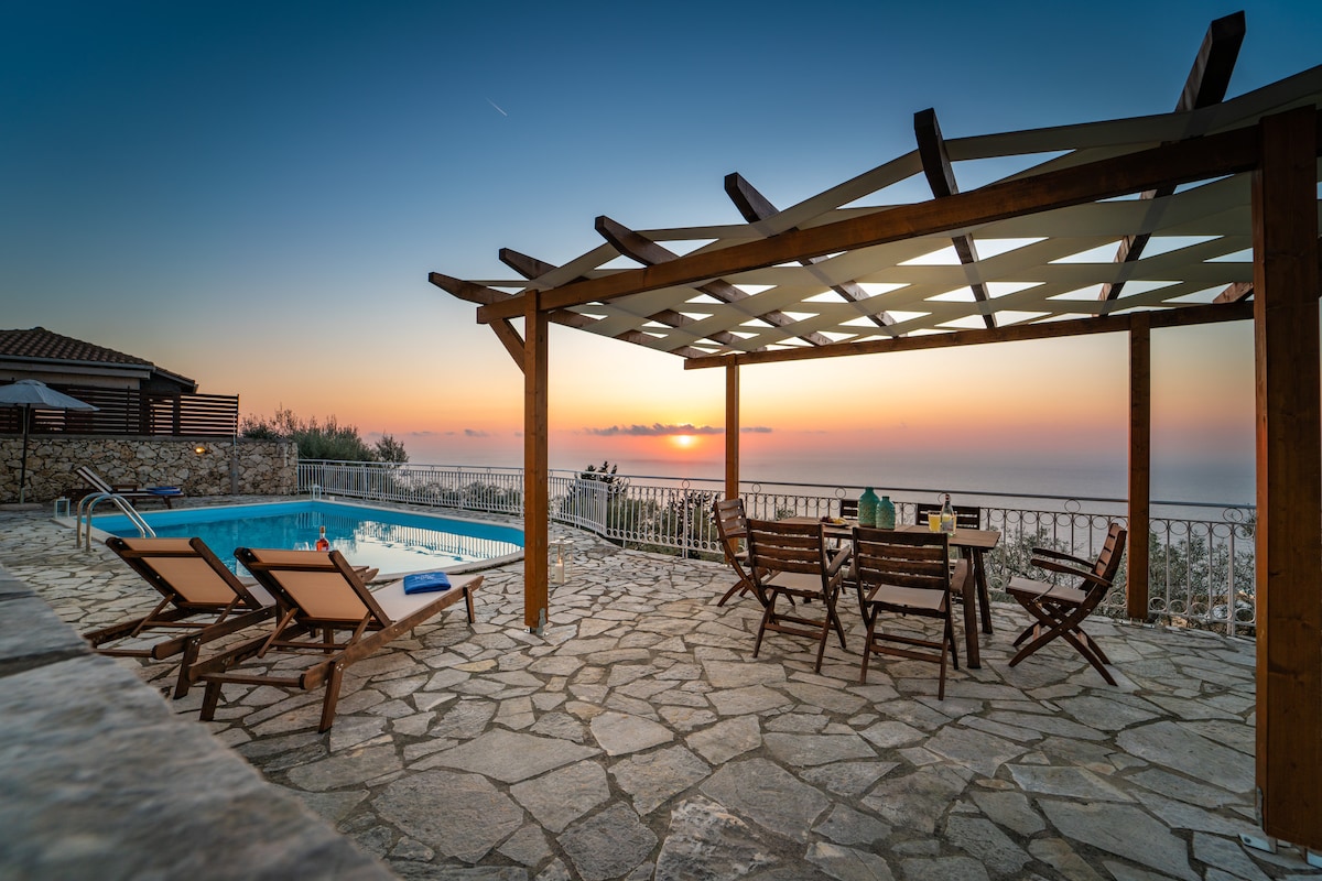 An outdoor patio area features a rectangular pool and several lounge chairs arranged around it. A wooden table with matching chairs sits under a pergola, providing shade. The backdrop showcases a sunset over the Ionian Sea, enhancing the natural beauty of the setting.