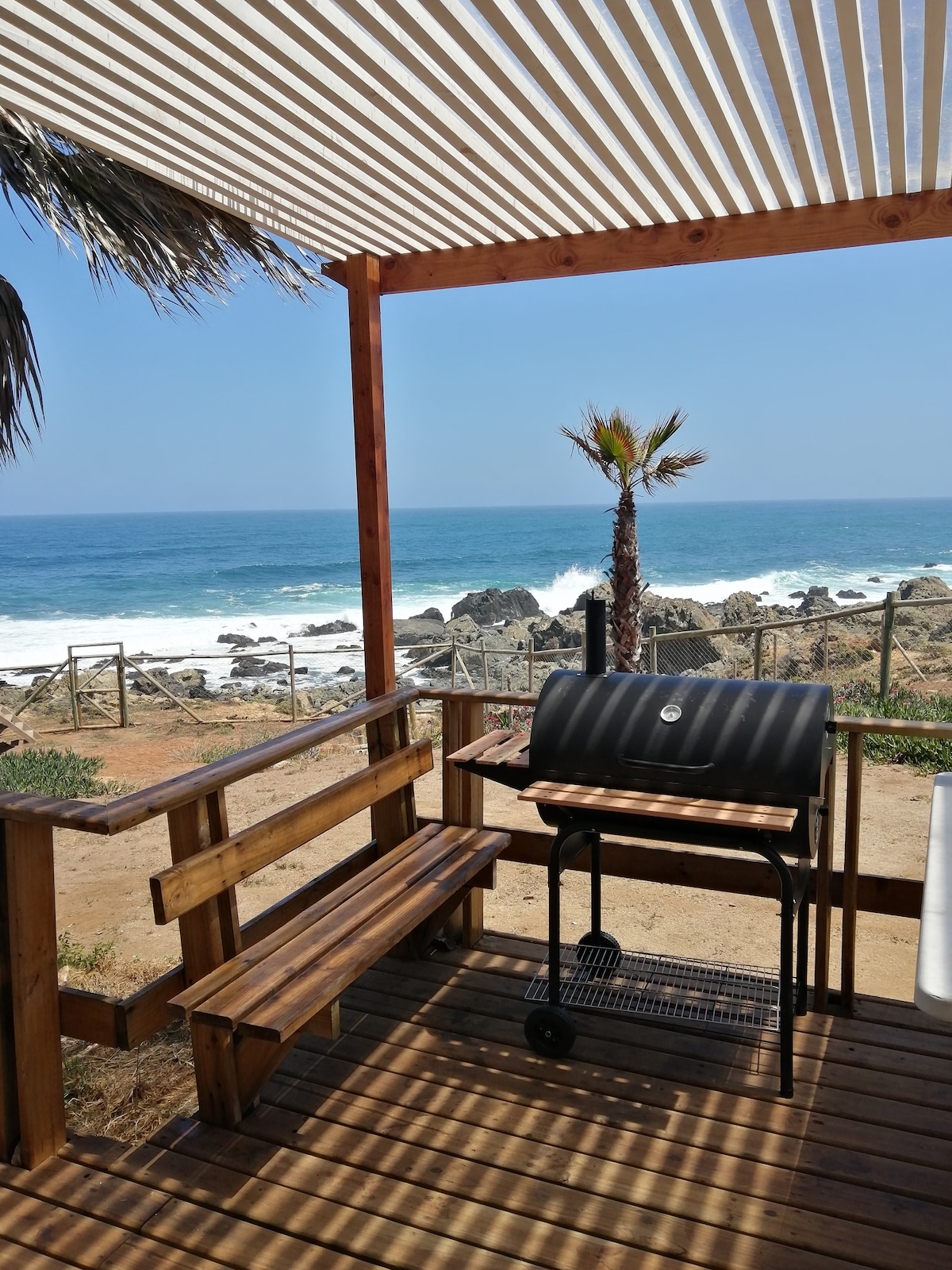 A shaded outdoor deck features a wooden bench and a charcoal grill, positioned to overlook the rocky coastline. Palm trees are visible, and the rhythmic waves of the ocean create a serene backdrop. Sunlight filters through the slatted roof, enhancing the peaceful setting.