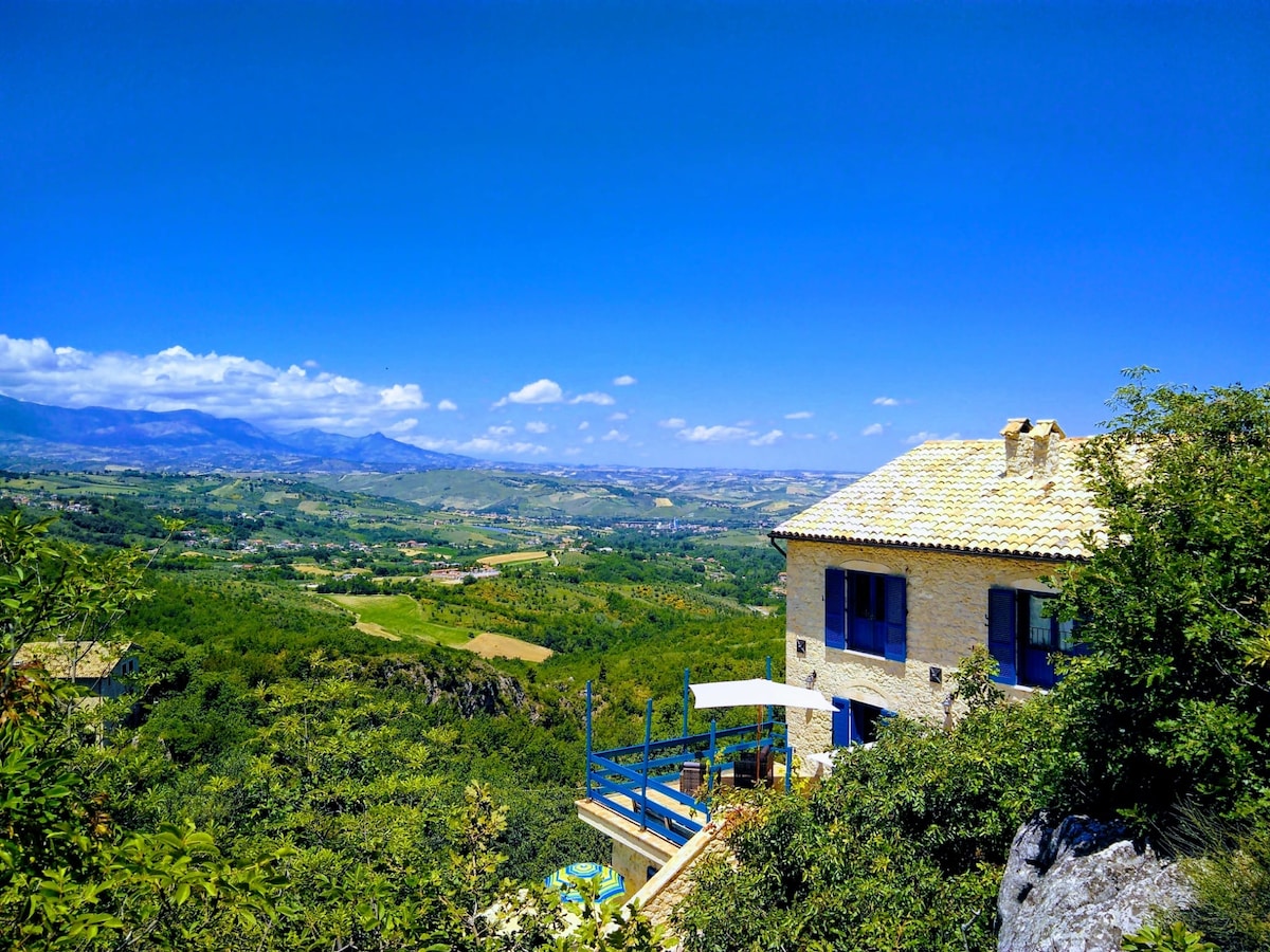 A rustic house with blue windows overlooks a lush green valley surrounded by rolling hills. A patio area with outdoor seating is visible in the foreground, with a clear blue sky and distant mountains enhancing the serene landscape.