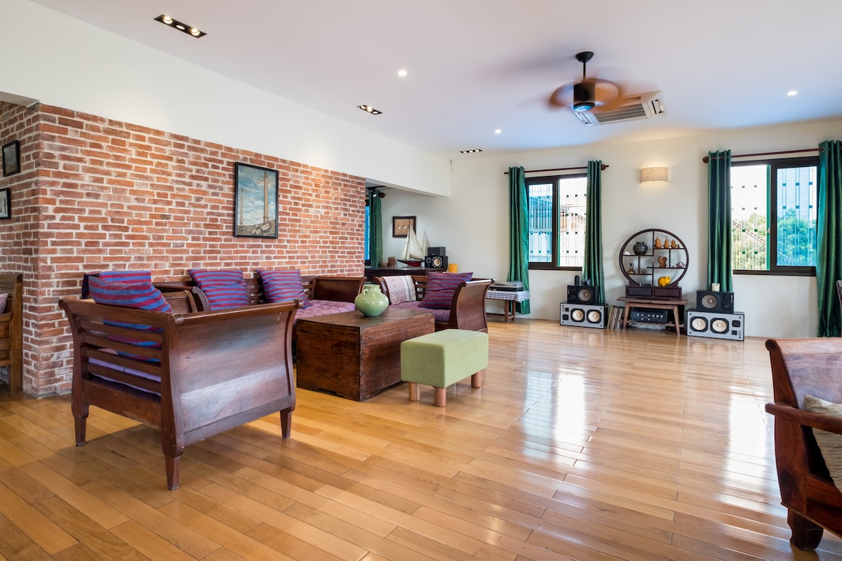 A spacious living area features wooden flooring and exposed brick walls, creating a warm ambiance. Comfortable seating arrangements include wooden chairs with colorful cushions. Large windows allow natural light to illuminate the room, with green curtains enhancing the décor.