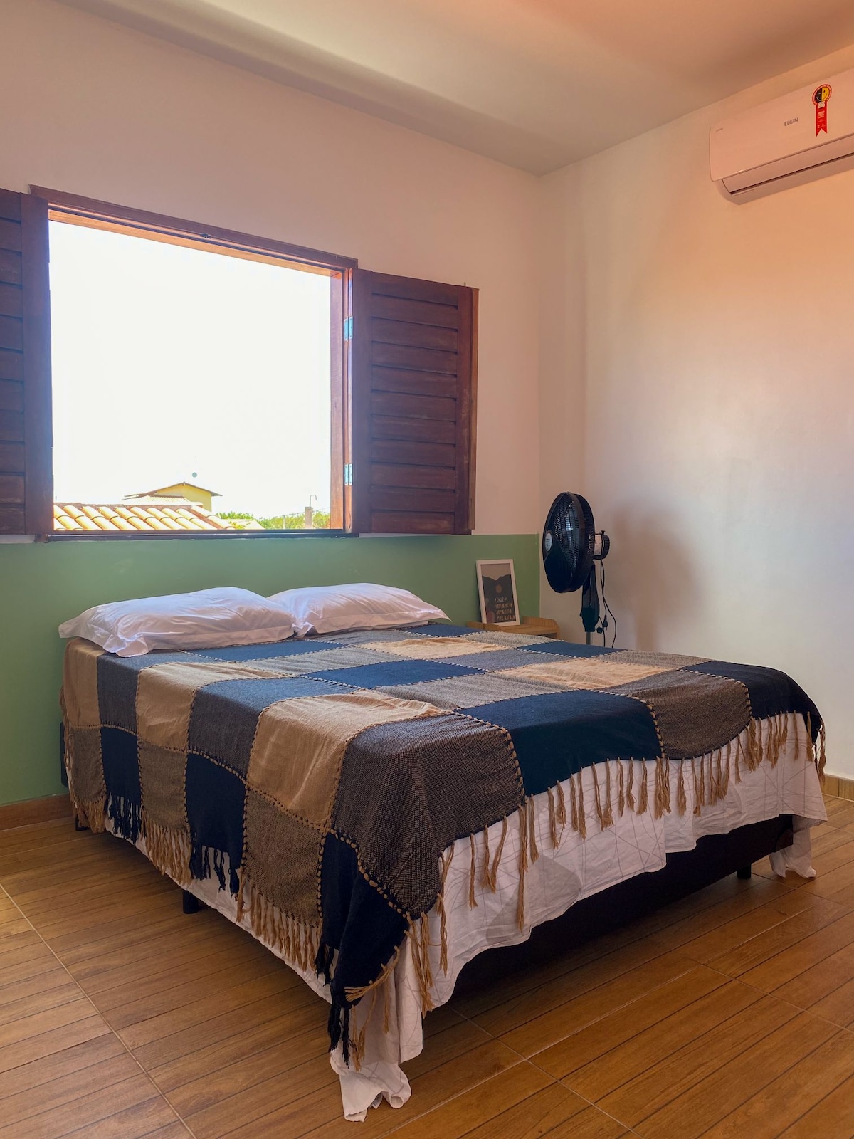 A cozy sleeping area features a spacious bed adorned with a patterned blanket. Natural light enters through a large window with wooden shutters, illuminating the room. A fan is positioned nearby, providing additional comfort in the warm space.