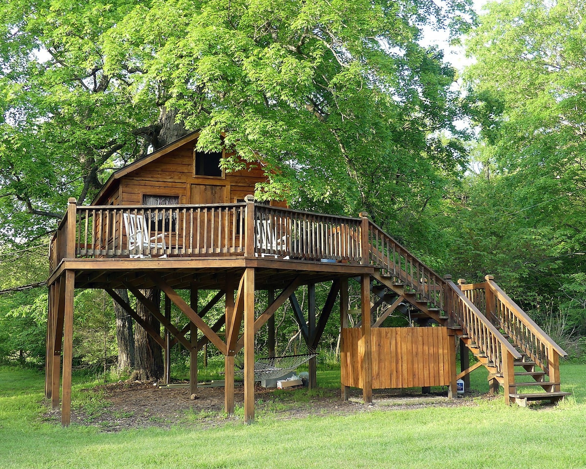 A rustic treehouse is elevated on wooden stilts, surrounded by lush green trees. A spacious deck features several outdoor chairs for relaxation. Steps lead up to the entrance, while a cozy hammock is nestled beneath. The landscape includes a grassy area encouraging outdoor activities.