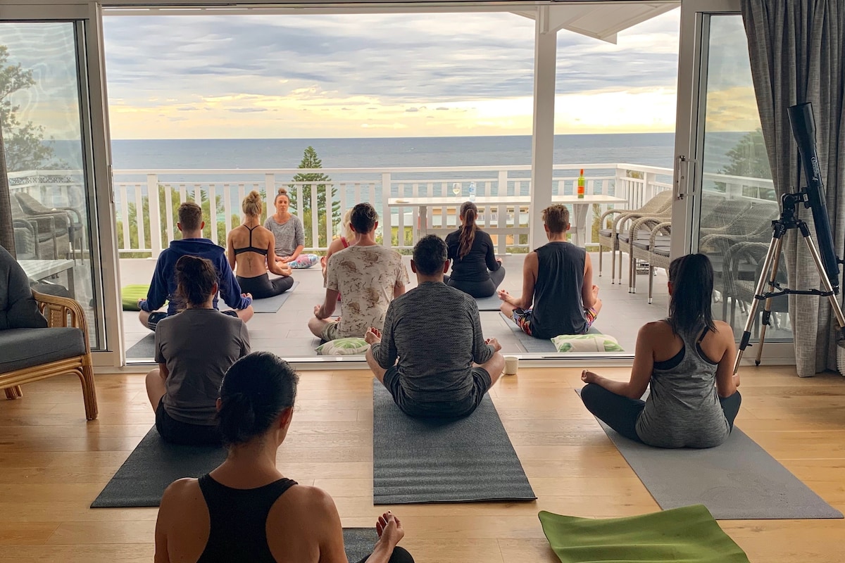 A group of individuals is seated on yoga mats, facing a large glass door that offers sweeping views of the ocean. The serene scene features gentle clouds in the sky, hinting at the calm atmosphere. Indoor plants and a telescope are also visibly placed nearby.