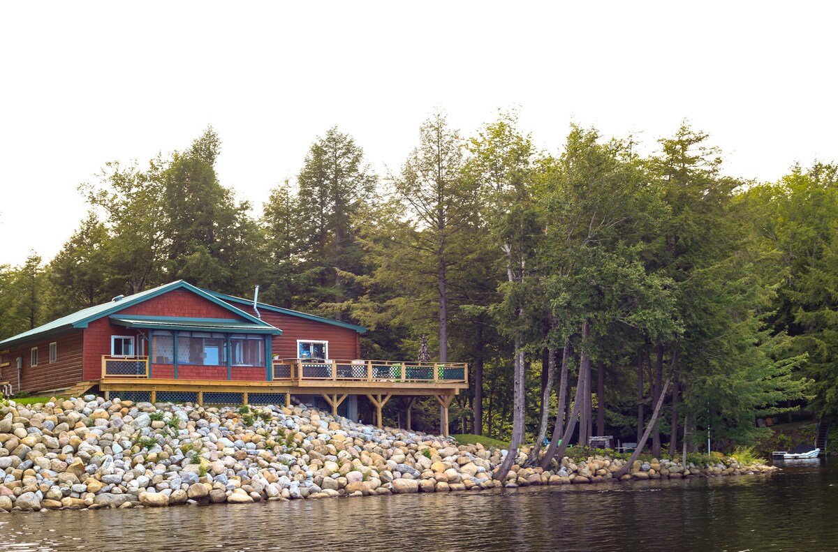 A lakeside house features a wooden deck that extends toward the water, surrounded by a pebble shoreline. The structure has large windows and a welcoming front entry. Trees provide natural privacy, while the serene lake reflects the calm surroundings.